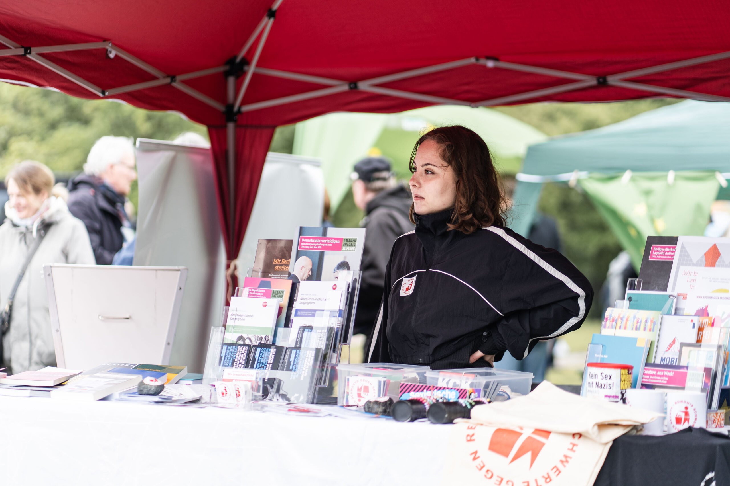 Ein Mitglied des Bündnis Schwerte gegen Rechts steht unter einem roten Pavillion. Vor der Person liegt auf einem Tisch ganz viel Infomaterial, es stehen Boxen mit Aufklebern und liegen Bündnis-Taschen aus. Im Hintergrund sind viele Menschen und weitere Stände zusehen.