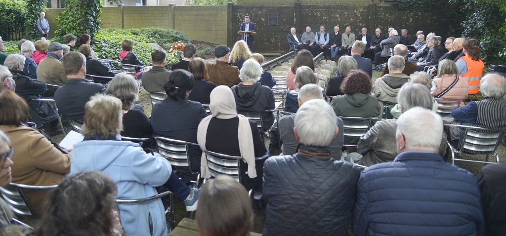 Auf dem Geländes ehemaligen KZ-Außenlager Buchenwald sitzen viele Menschen um die Schienen mit Figuren von Menschen als Schwellen, angebracht als Mahnmal zur Erinnerung an das ehemalige KZ. ie schaueb zu einem Redepult an dem Bürgermeister Dimitrios Axourgos eine Rede hält.