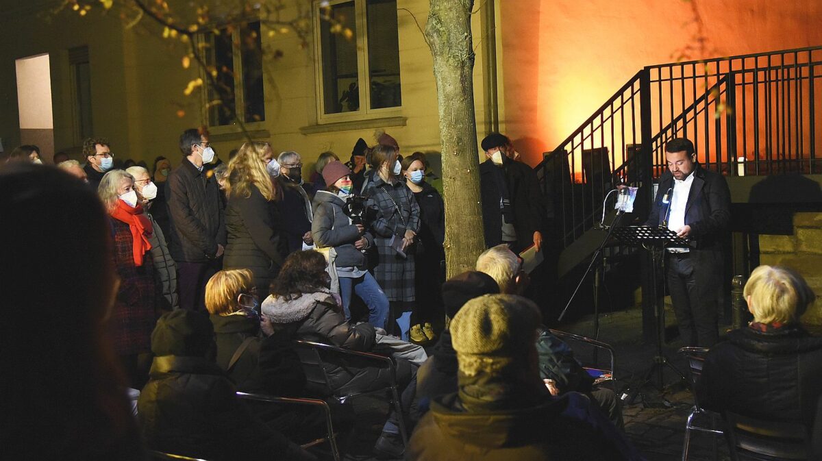 Mehre Menschen stehen und sitzen im dunkeln um der alten Synagoge vor der ein Rednerpult steht, an dem der Bürgermeister eine Rede hält.