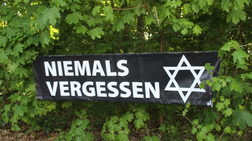 Zwischen zwei Bäumen hängt ein Banner mit Davidsterne und der Aufschrift "Niemals Vergessen".
