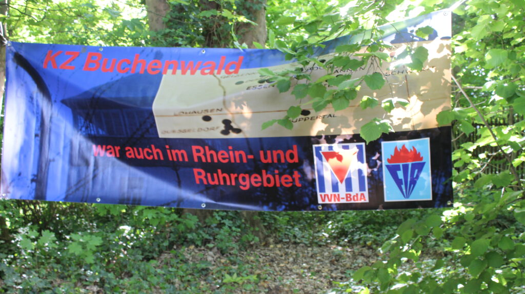 Zwischen zwei Bäumen hängt ein Banner vom VVN BDA mit der Aufschrift "KZ Bucenwald war auch im Rhein- und Ruhrgebiet".