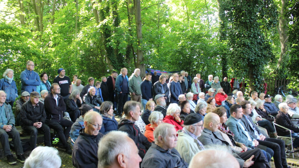 Ca. 50-100 Menschen sitzen und stehen auf dem Geländer der Gedenkstätte.